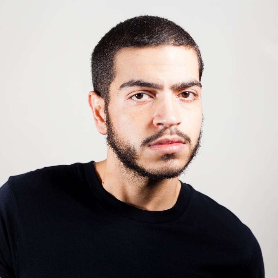 Bearded Puerto Rican gay male shoulders portrait looking straight to the camera handsome, serious, honest and confident with short hair and black shirt on a soft grey background. Male sight expression