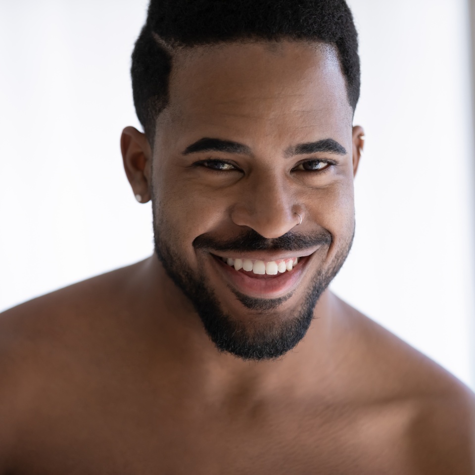 Close up portrait of attractive African American guy with stylish beard, piercing, white healthy teeth, athletic shoulders looking at camera and smiling. Male beauty care concept. Head shot