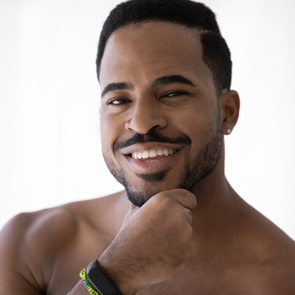 Portrait of handsome African American man with stylish beard touching stubble, applying aftershave balm, looking at camera with toothy smile. Metrosexual, male beauty care concept. Head shot