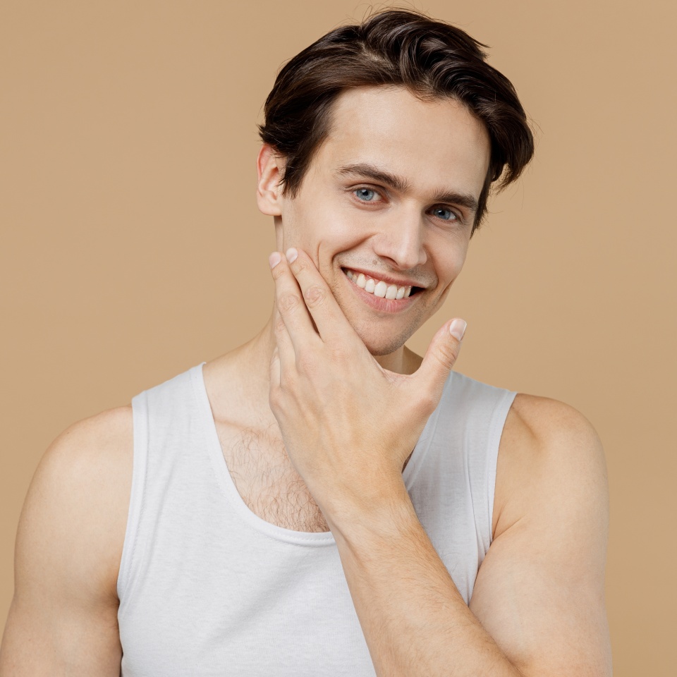 Attractive smiling calm young man he 20s perfect skin wearing bundershirt touch face after shaving isolated on light pastel beige background studio. Skin care healthcare cosmetic procedures concept.