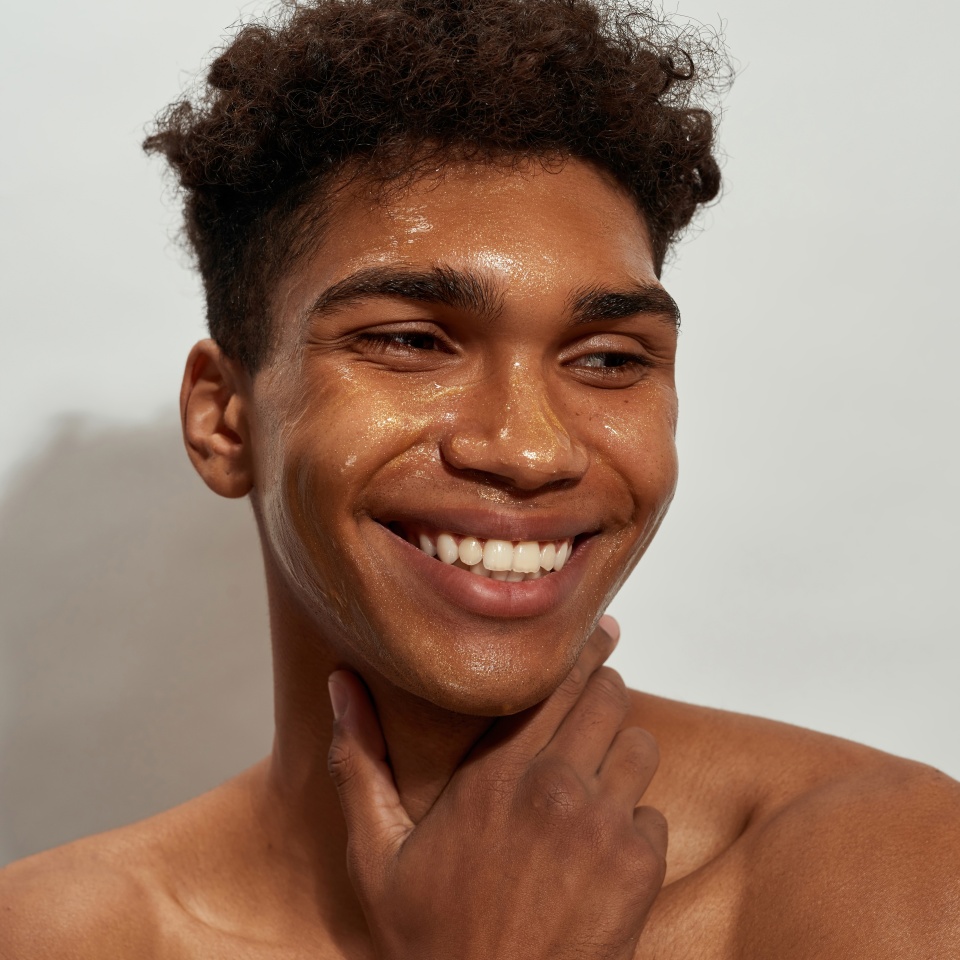 Portrait of smiling black guy with golden mask on his face looking away. Young slim brunette curly man with naked torso. Facial skin care. Isolated on white background. Studio shoot