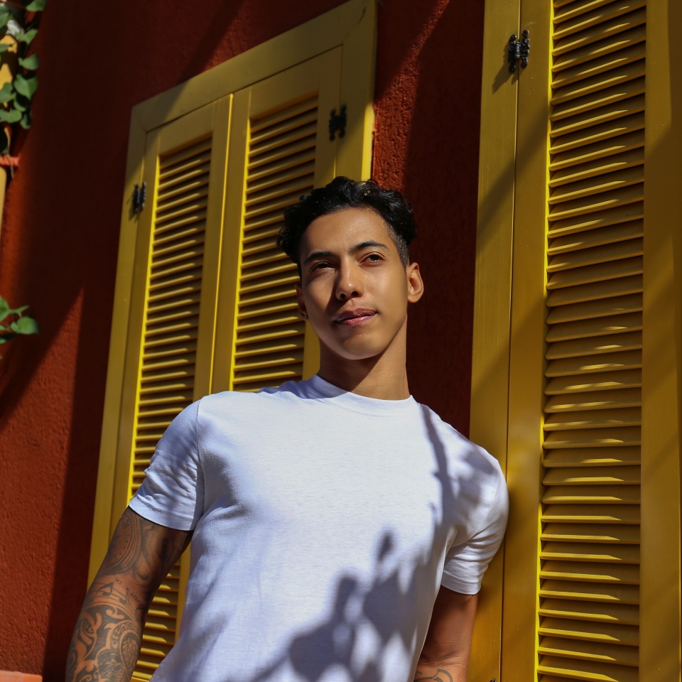 Handsome young latin man in plain white t-shirt standing near the rustic house with red walls and yellow window shutters. Close up, copy space, background.