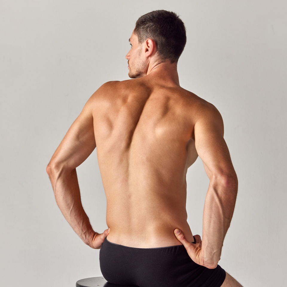 Back view image of muscular young shirtless man with relief, sign back sitting in underwear against grey studio background. Concept of men's health and beauty, body care, fitness, wellness, ad