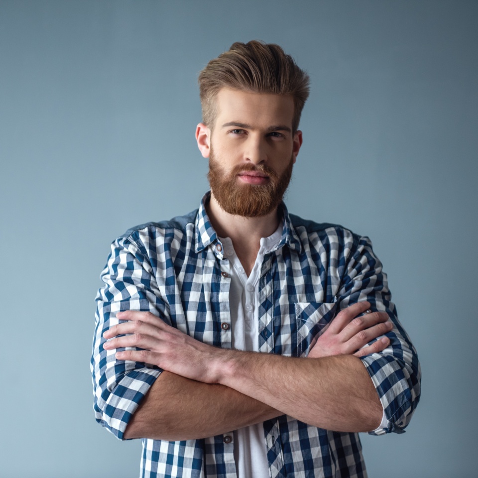 Handsome bearded man in casual clothes is looking at camera while standing with folded arms on gray background