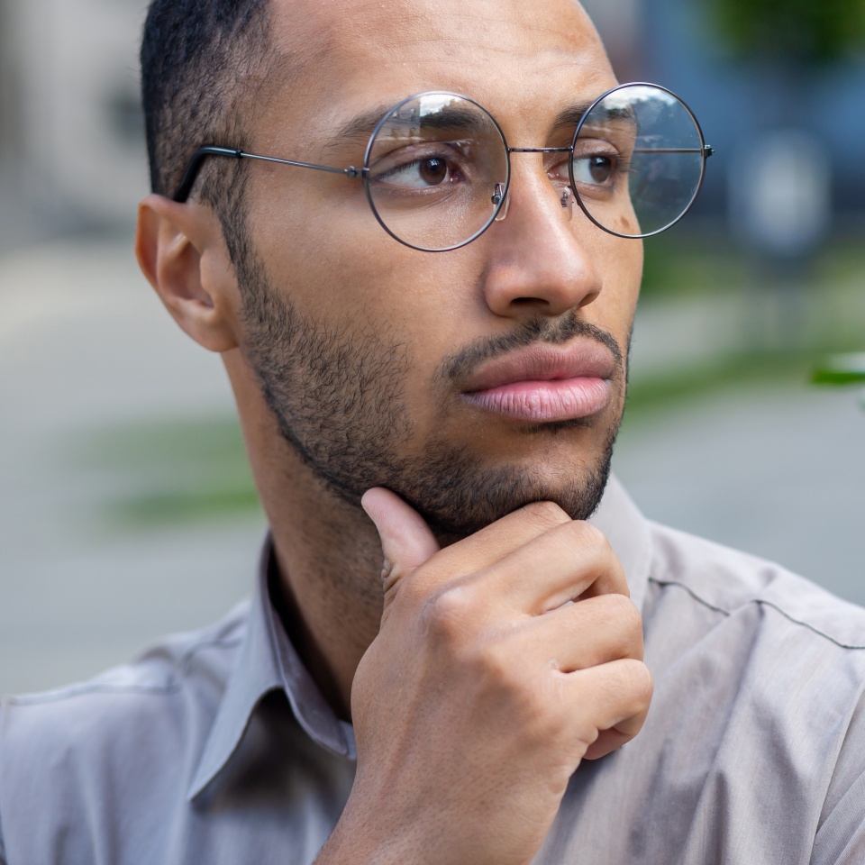 A close-up photo of a young Hispanic man standing outside the city wearing glasses, rubbing his beard with his hand, looking seriously and thoughtfully to the side.