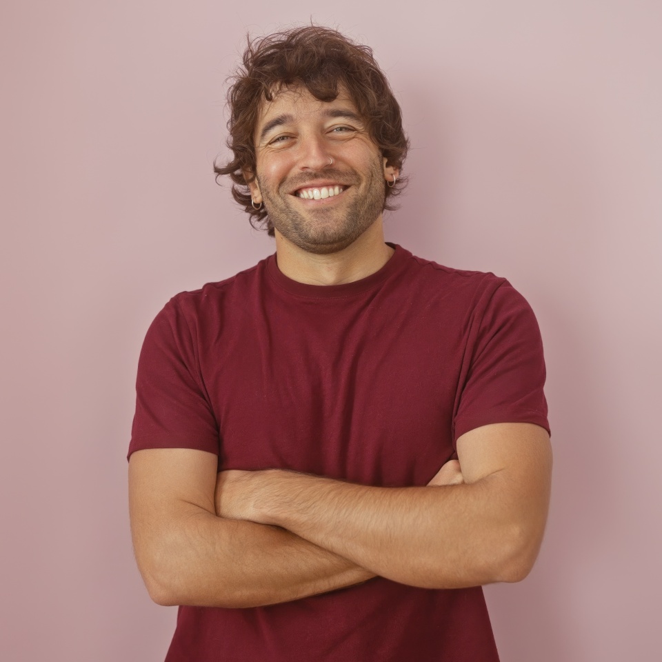 Handsome hispanic man with a beard smiling with arms crossed against a pink wall in a casual maroon shirt.