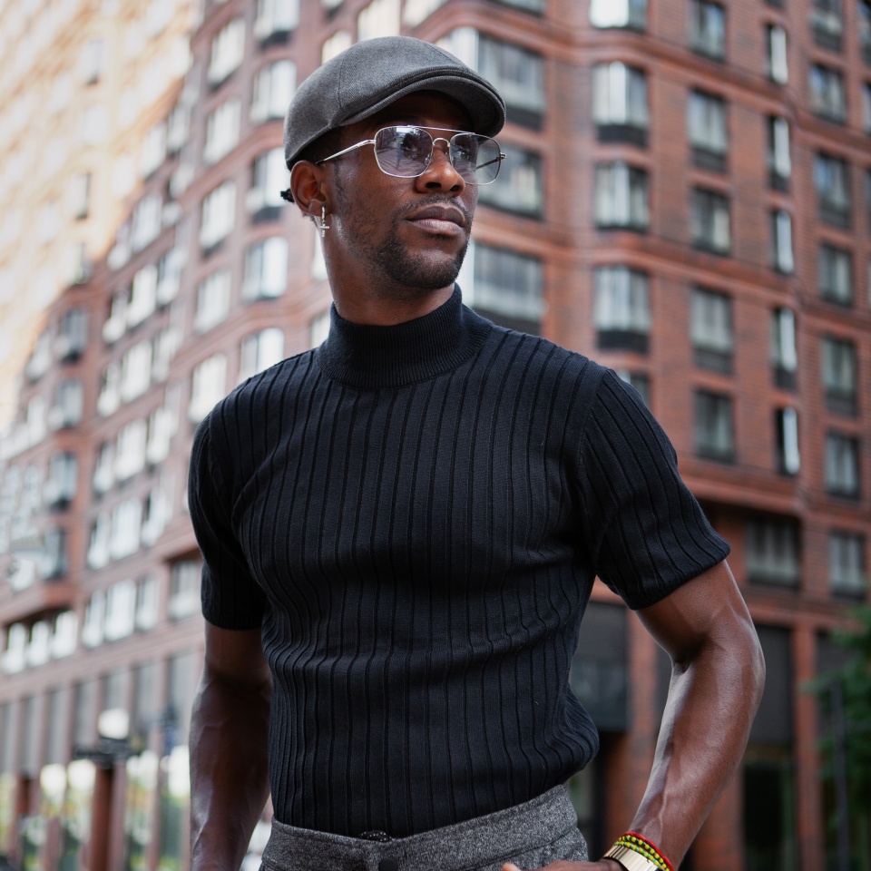 Handsome stylish African American man stands on the street in fashionable attire, wearing a cap, sunglasses, and a black t-shirt, modern male fashion model. Trending men's style of clothing.