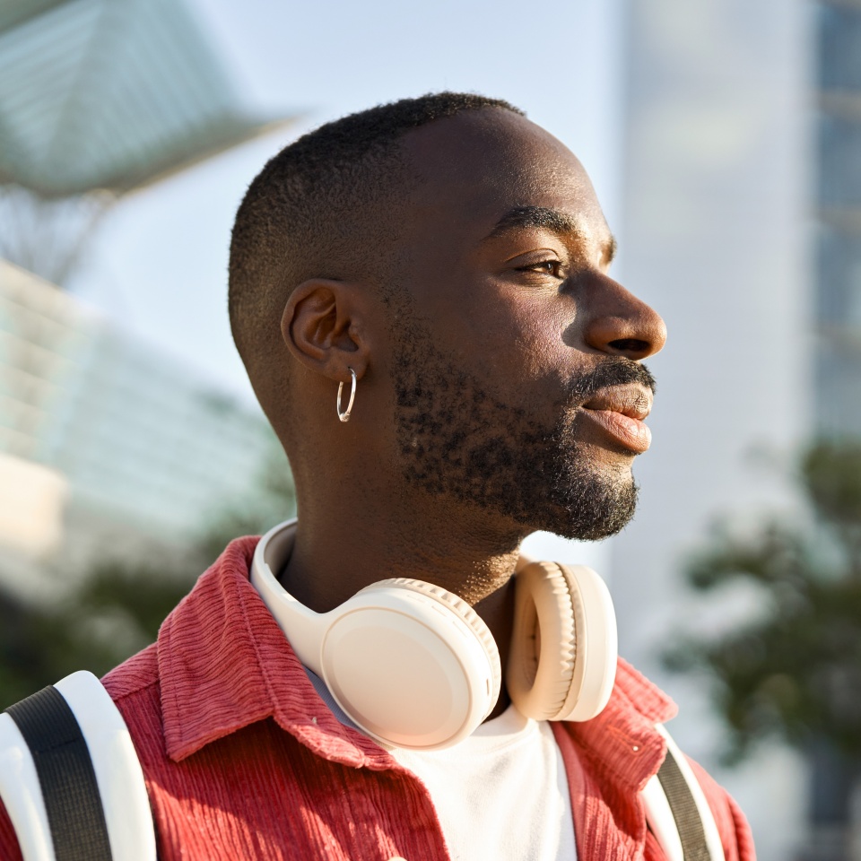Portrait of happy young adult African man hipster standing on city street outdoors looking away dreaming. Smiling cool gen z Black hipster guy traveler wearing red shirt headphones backpack. Close up