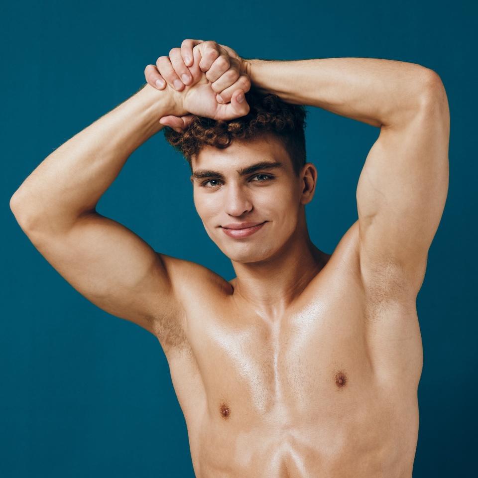 Confident young man posing with muscular arms raised, showcasing strength and vitality against a teal background. Self-expression and fitness concept.