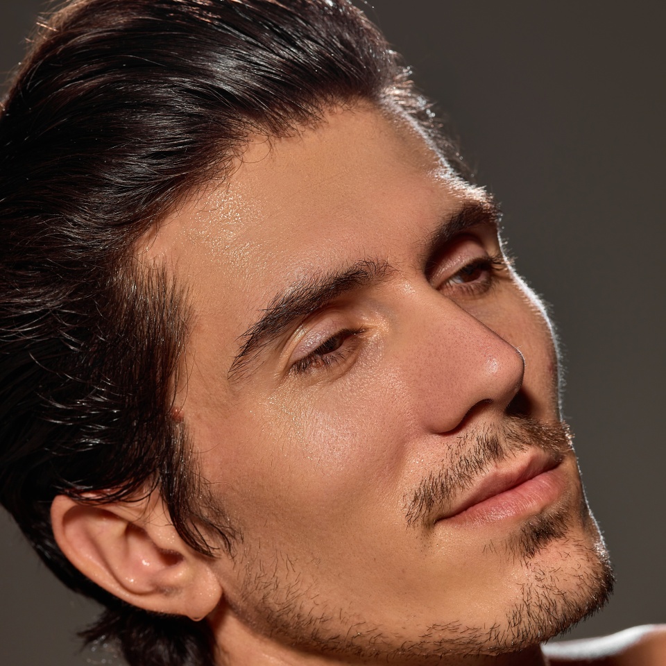 Close-up of man's face with head tilted back against dark background. Skin with natural glow, showing smooth and clear texture. Hair styled back neatly. Concept of male beauty and facial care,