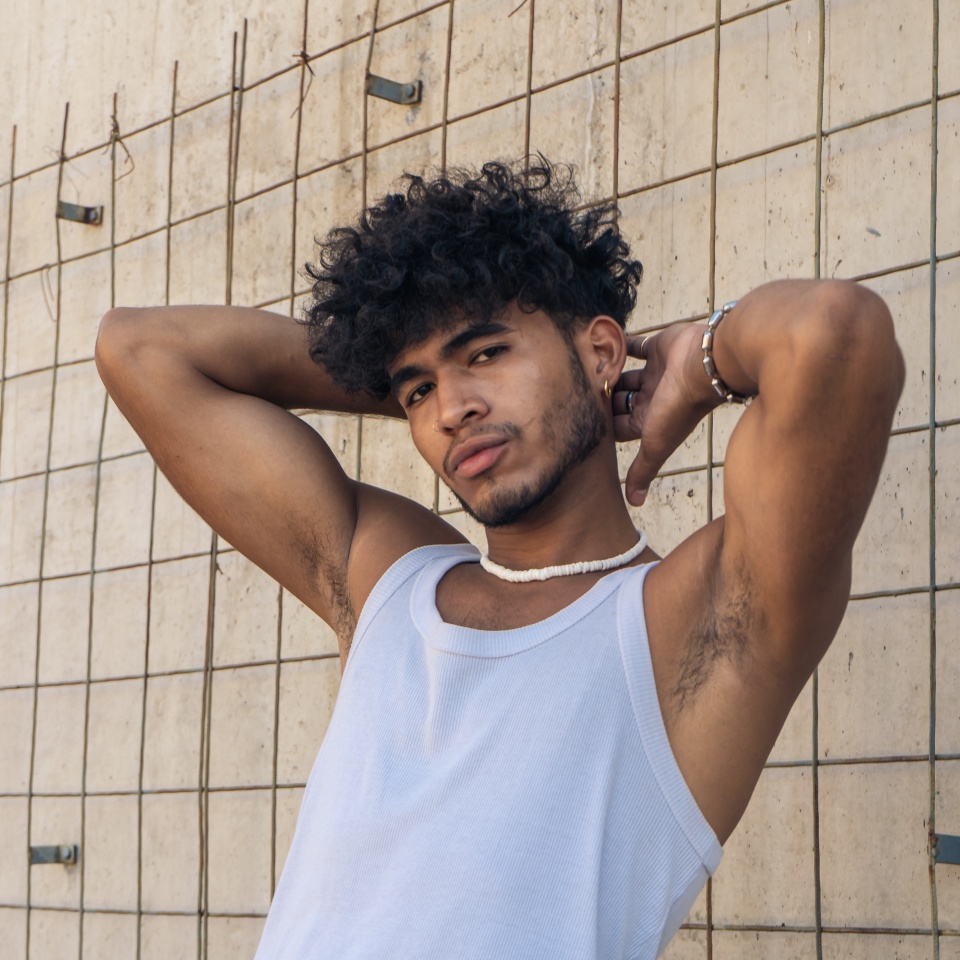 A handsome young Latino man with a personal style sitting on a bench in front of a large cement wall.