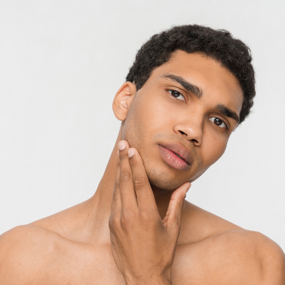 Photo of attractive black man african american guy touches his smooth young clean fresh face and looks at camera after shaving lotion result. Naked torso, isolated white color background