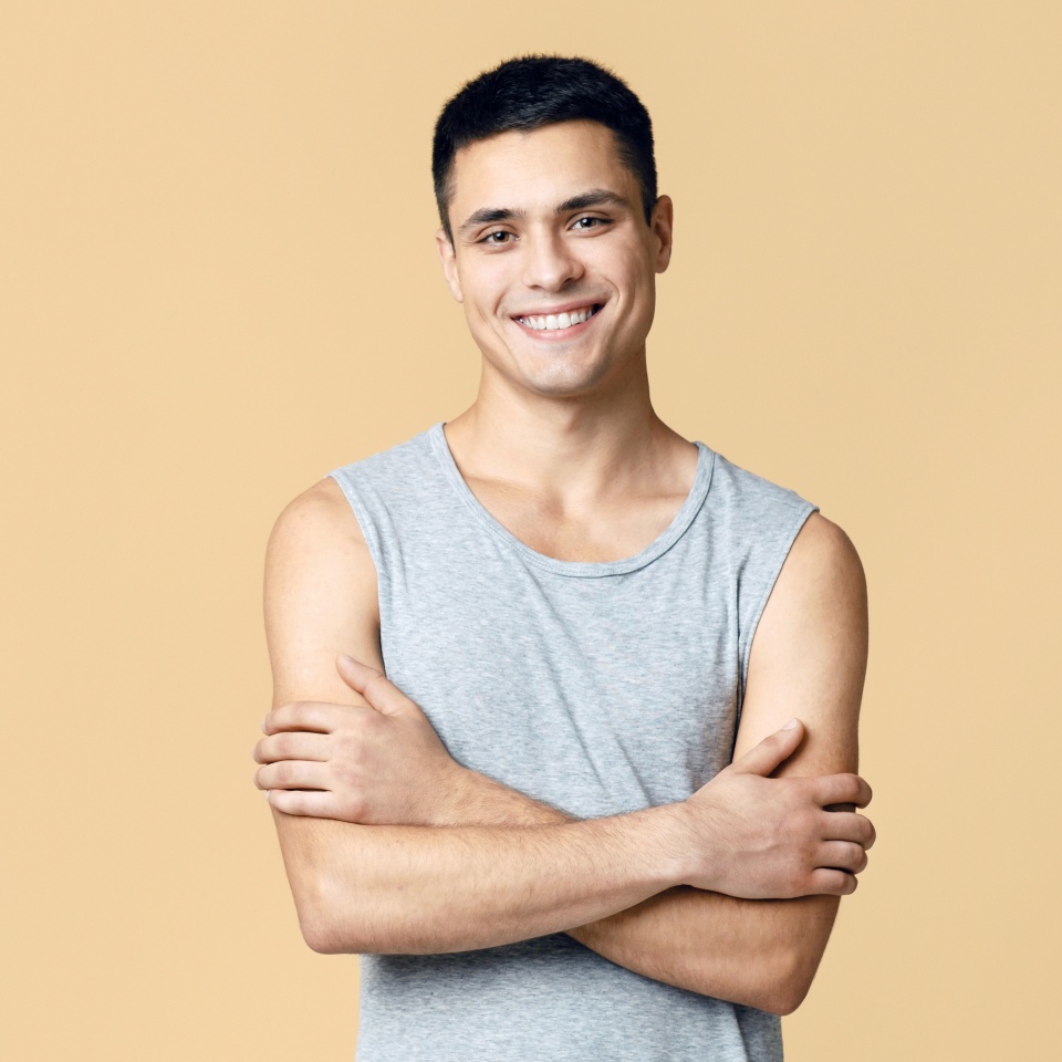 Handsome attractive muscular guy model or blogger and real emotions. Smiling friendly man in casual with crossed arms looking at camera, isolated on light background, empty space, studio shot