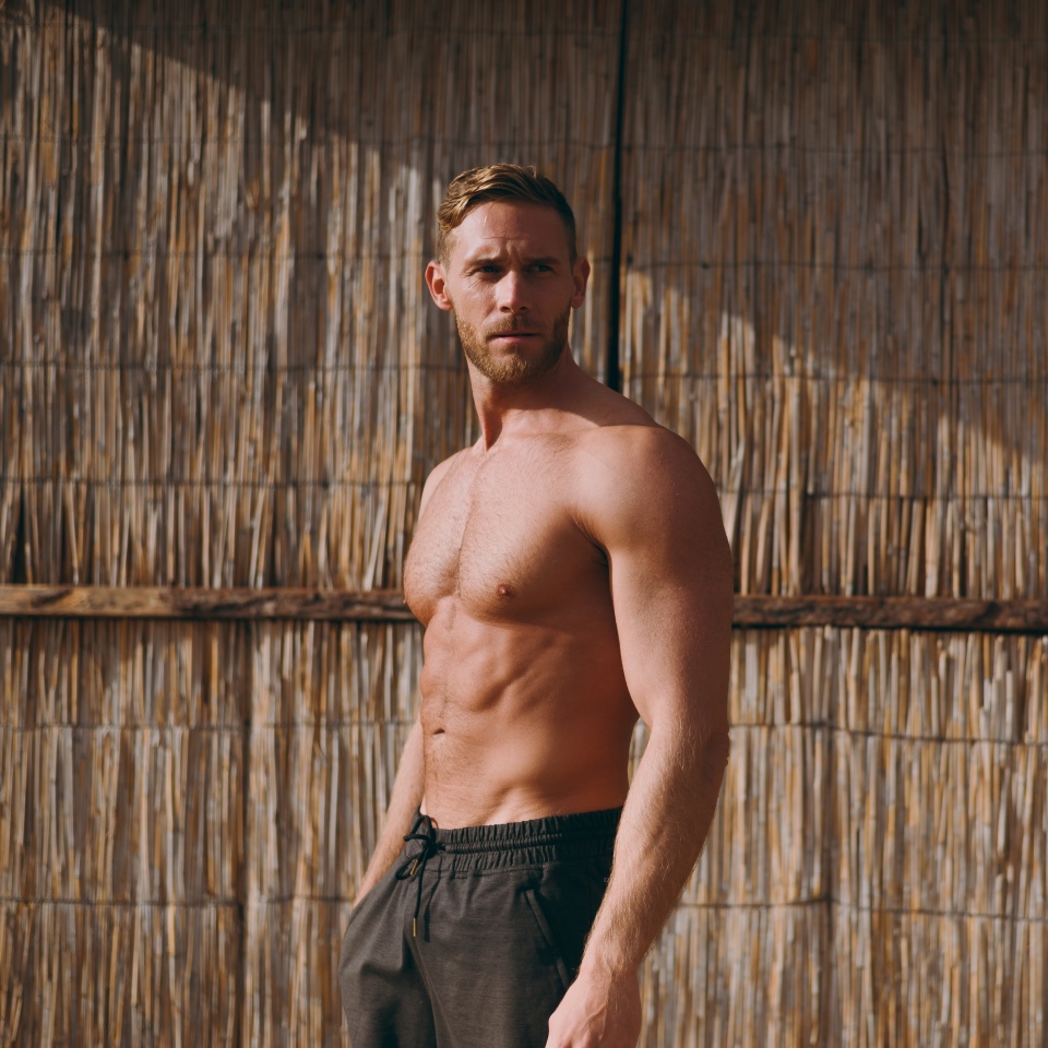 Side profile view young strong sporty athletic toned fit sportsman man wear shorts with naked torso look aside stand near straw wall after warm up training on sea beach outdoor in summer day morning