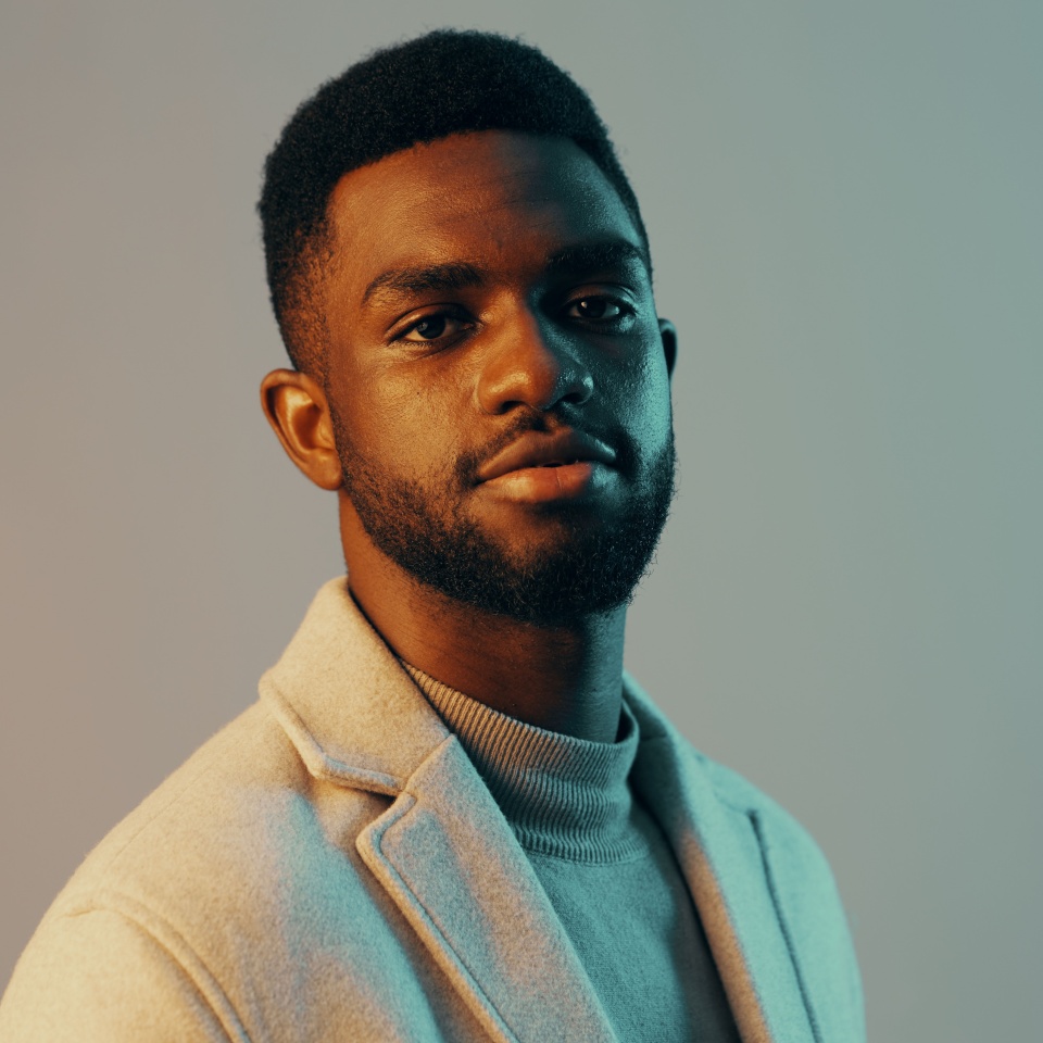 Portrait of a stylish young Black man with a confident expression, wearing a light grey coat and sweater, set against a soft color gradient background