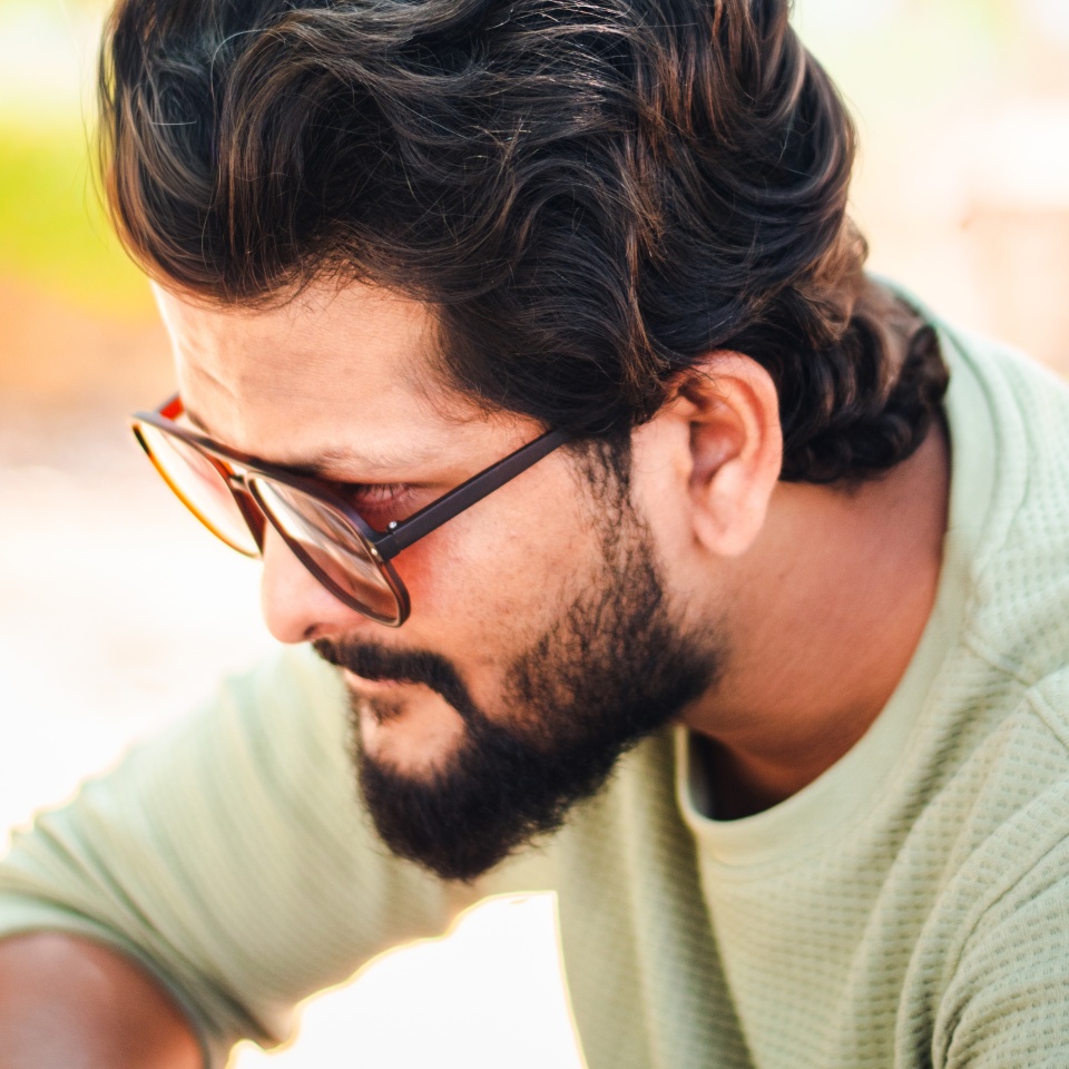 Close-up portrait of a young Indian man with wavy hair and beard, wearing glasses, outdoors in India.