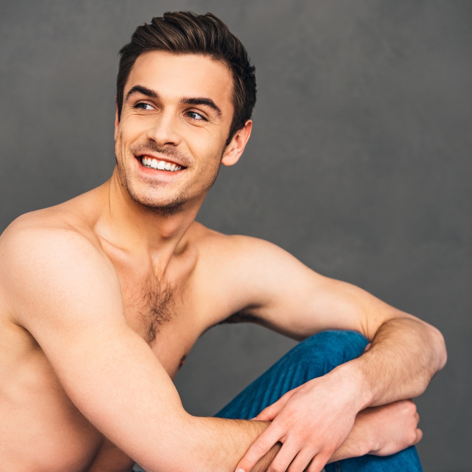 Feeling happy. Cheerful handsome young man looking away with smile while sitting on bed against grey background