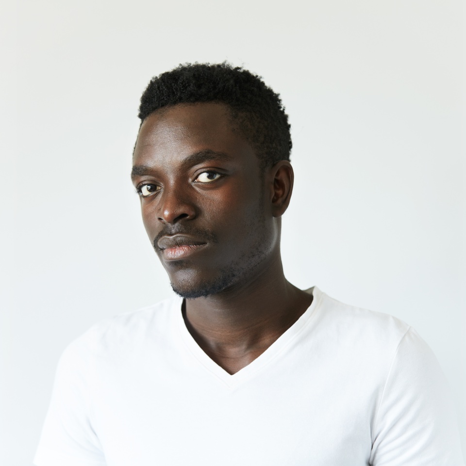 Headshot of handsome young African man wearing white T-shirt, looking at the camera, posing isolated against white concrete wall background with copy space for your information or advertising content
